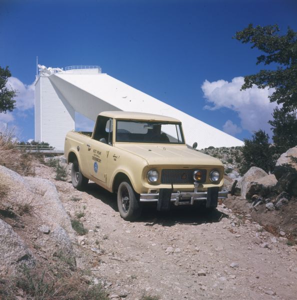Person driving a yellow Scout pickup. "Kitt Peak National Observatory" is painted on the passenger door. In the background is the observatory building.
