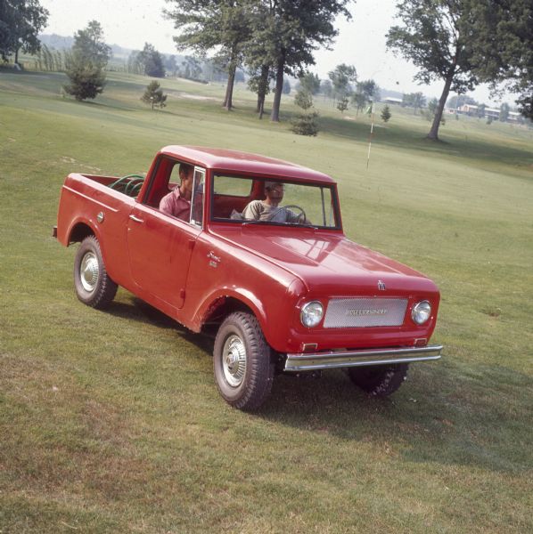 Two men in the cab of a red Scout pickup. They are driving on a golf course, and a green hose is visible in the back of the open pickup.