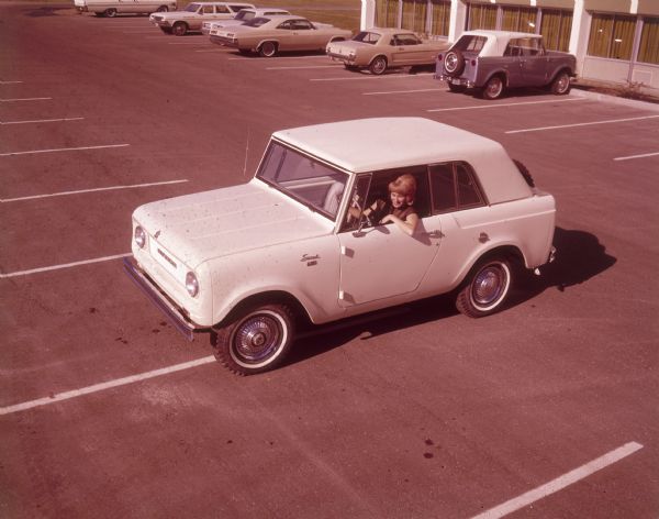 Elevated view of woman driving a white Scout in a parking lot. A spare tire is mounted outside on the back of the truck. Cars are parked in the background near a building.