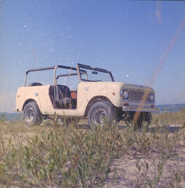 Three-quarter view from front of passenger side of Scout parked on beach. The Scout has a windshield and roll bars in place, but no doors. The ocean is in the background.