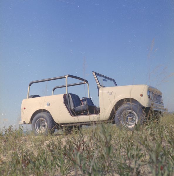 Passenger side view of Scout parked on dunes. The Scout has a windshield and roll bars in place, but no doors. The ocean is in the background.