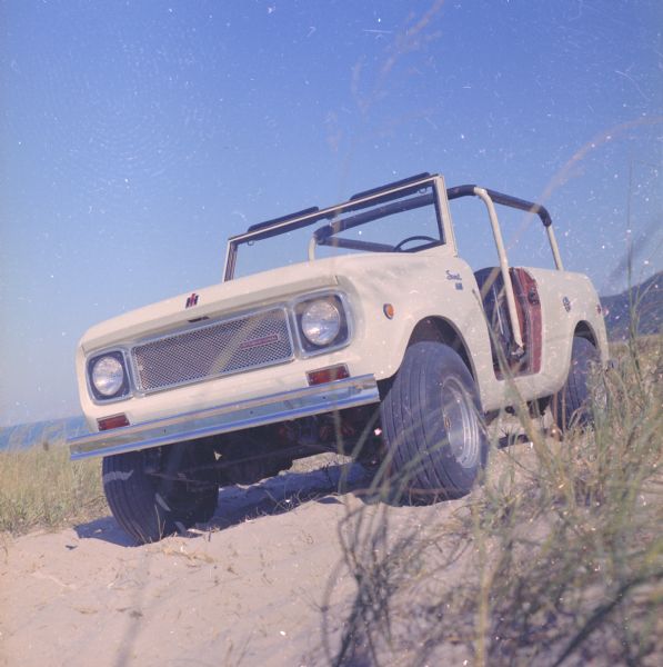Three-quarter view from front of driver's side of Scout parked on beach. The Scout has a windshield and roll bars in place, but no doors. The ocean is in the background.