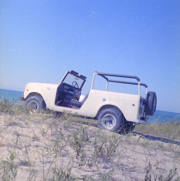 Driver's side view of Scout parked on beach. The Scout has a windshield and roll bars in place, but no doors. A white helmet is sitting on the driver's seat. The ocean is in the background.