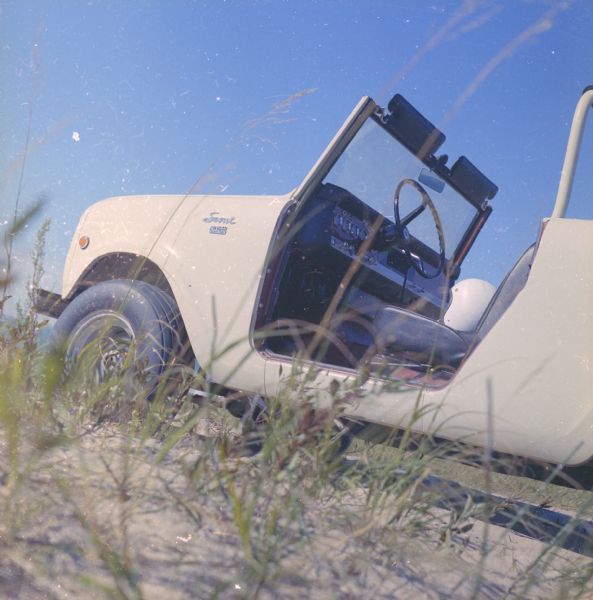 Close-up of driver's side of Scout parked on dunes. The Scout has a windshield and roll bars in place, but no doors. A white helmet is sitting on the driver's seat. The ocean is in the background.