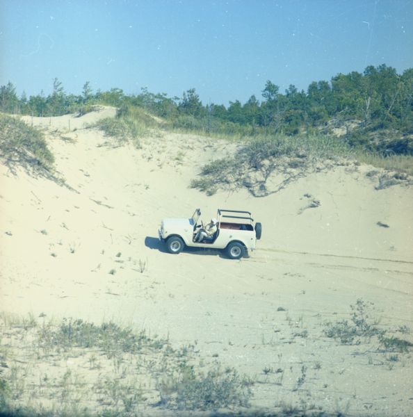 View looking down dune towards a man wearing a helmet driving a Scout. Trees and plants are on top of the dunes. The Scout has a windshield and roll bars in place, but no doors.