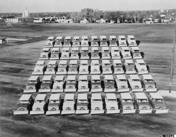 Elevated view of fifty Scout snow plows parked outdoors in a large field. There are factory buildings in the background.