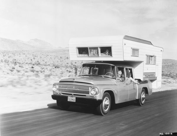 Man, woman, and a young child holding a pinwheel outside the window, are riding in an International 1966 Travelette pickup with camper attached. They are traveling on a road in a desert area. Camper is slide-in version. Two young children are at the front window of the camper, which is over the cab.