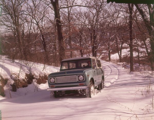 Three-quarter view from front of driver's side of International Scout with Traveltop. A man is driving on a snowy road, breaking new tracks in a hilly, wooded area.