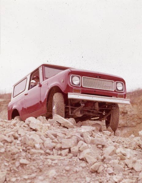 Three-quarter view from front of passenger side of red International Scout with white Traveltop. A man is looking out of the open passenger side window as the Scout is traveling up a rocky incline.