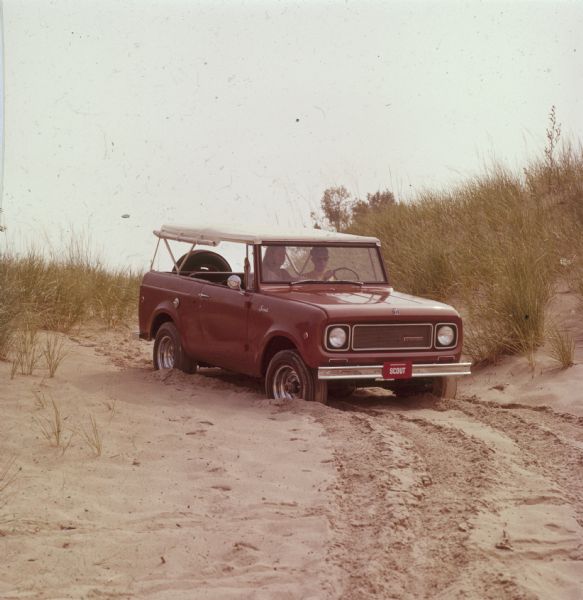 Three-quarter view from front of passenger side of red International Scout with white Traveltop. Two people are in the front seat. The sides of the Traveltop are rolled up, and a spare tire is mounted outside on the back of the tailgate. A man is driving the Scout in sand near grassy dunes.