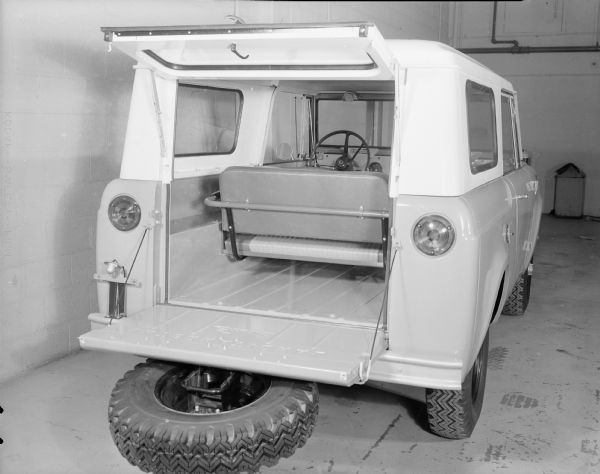 Rear view of Scout parked indoors. Tailgate is open and laying flat, with spare tire mounted on outside of tailgate. Window hatch of Traveltop is opened up towards roof. Rear seat is one long bench.