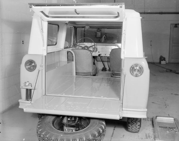 Rear view of Scout parked indoors. Tailgate is open and laying flat, with spare tire mounted on outside of tailgate. Window hatch of Traveltop is opened up towards roof.