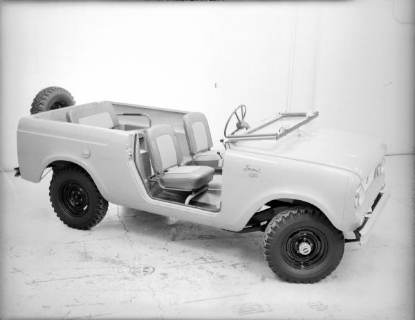 View of passenger side of Scout 4x4. The doors are removed and the windshield is folded down over the hood. Spare tire mounted on outside of tailgate. There is a rear bench seat in the open bed.
