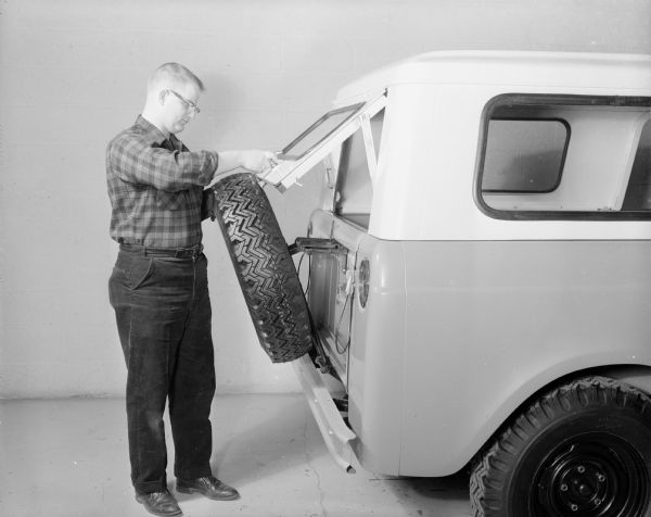 Side view of man standing behind the tailgate of a Scout 4x4 with Traveltop. He is tilting back the spare tire attached to the back of the tailgate with his left hand in order for the upper window hatch to be lifted up with his right hand.