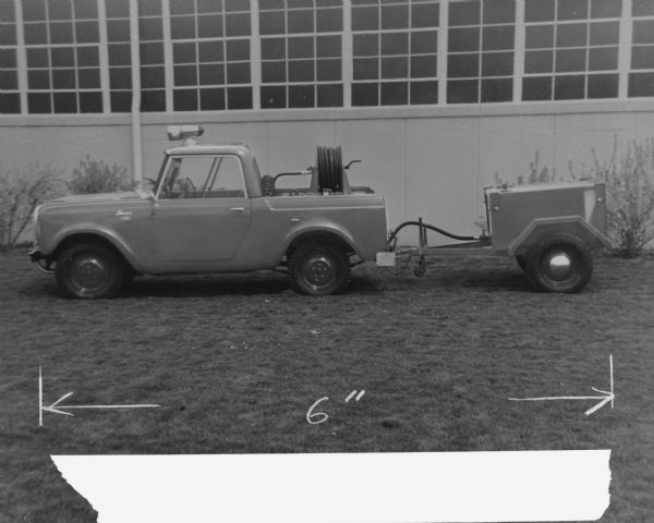 Side view of driver's side of Scout 4x4, pulling a trailer. Spare tire is mounted in truck bed on back of cab. Mounted in the trailer bed is a hose on a reel, and a hose is connected to the trailer. A search light is mounted on top of the cab. The truck and trailer are parked on a lawn in front of an industrial building.