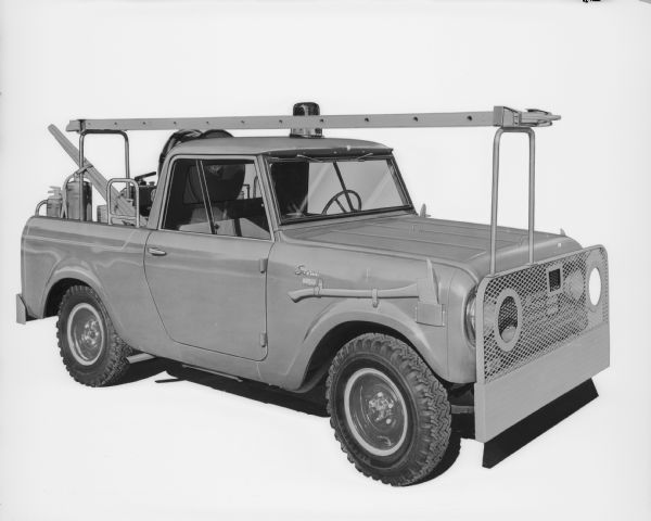 Three-quarter view from front of passenger side of Scout 4x4. Equipped with a ladder above cab on supports on front grille and in truck bed. Hose on reel in truck bed. Other equipment is in the truck bed, and a search light is on the roof of the cab.