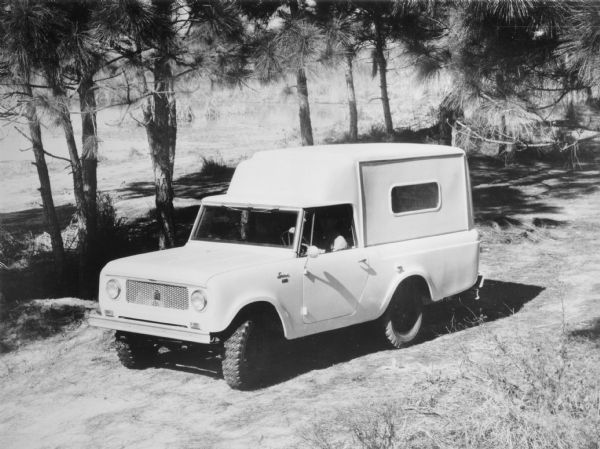 Slightly elevated three-quarter view from front of driver's side of Scout. The Scout has a large truck bed topper that rises higher than the cab roof. A man is in the driver's seat. There are pine trees and a pond or river in the background.