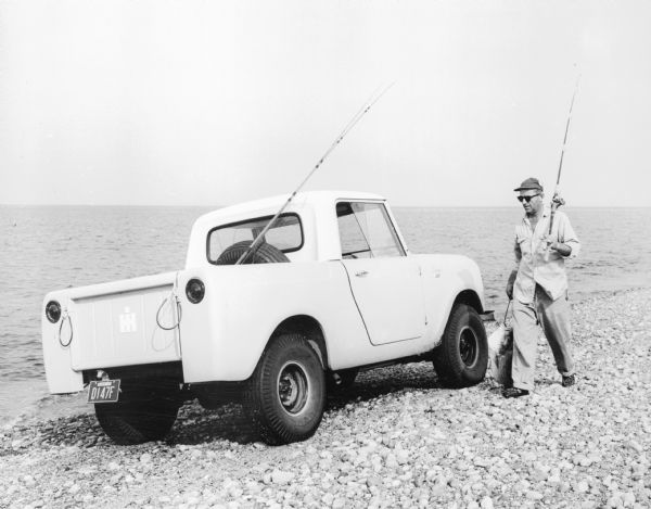 View of Scout parked on rocky shoreline. A man is walking near the Scout carrying a fishing pole and a large fish on a stringer. A fishing pole is sticking out of the truck bed where it rests against the spare tire mounted to the back of the cab in the truck bed.