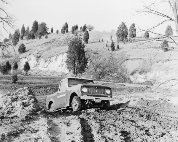 Three-quarter view from front of passenger side of Scout being driven on deep, muddy terrain. Painted on the passenger side door: "Pleasant View Farm." In the background is a hill and trees.