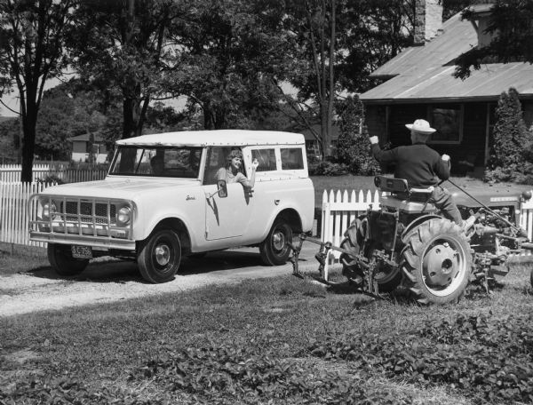 Woman driving a Scout with canvas topper along a driveway. She is wearing a string of pearls and is wearing gloves. A young boy is sitting in the passenger seat. In the background is a fenced-in yard and a house. A man driving a Farmall tractor near the driveway is waving to the woman and she is waving back.