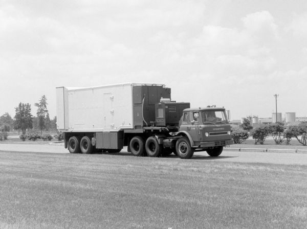 COF-1810A Cargostar with Missile-Launch Cell. A group of men are in the cab of the Cargostar which is traveling along a road. In the background are industrial buildings and storage tanks.