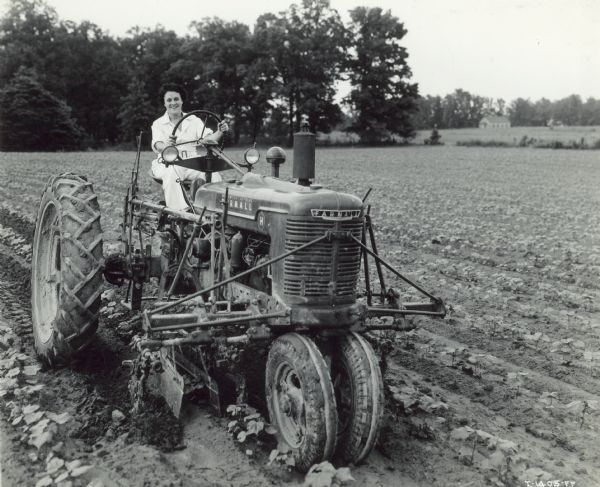 Three-quarter view from front left of a woman on a Farmall H tractor in a field. Caption on accompany photograph: "A very successful tractorette class of twenty women was organized by Hinton & Hutton, of Collierville, 23 miles east of Memphis, Tennessee. A prominent member of the class was Mrs. John T. Shea who is shown solo in cotton field on her 150-acre plantation in I-1405-FF, I-140-FF, and I-1407-FF and who regularly operates the tractor."