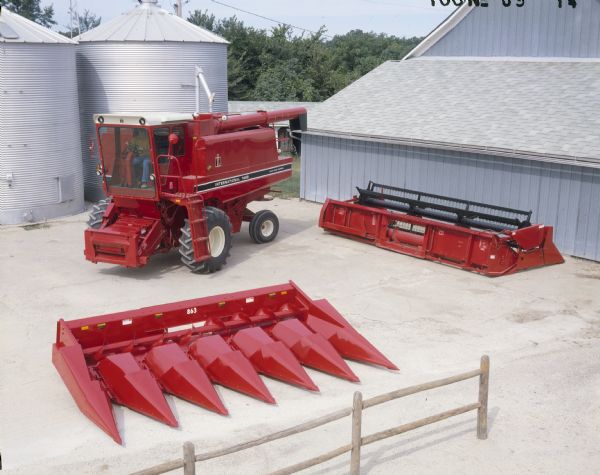 Elevated view of a man sitting in a 1460 axial-flow combine. On the ground nearby is a 6 row corn head on the left, and on the right is a grain head. A farm building and two silver colored silos are behind the combine.