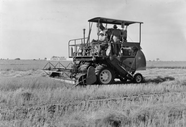 McCormick-Deering self propelled combine in flax. The driver and bagger are perched atop the machine. Both men are wearing hats. 