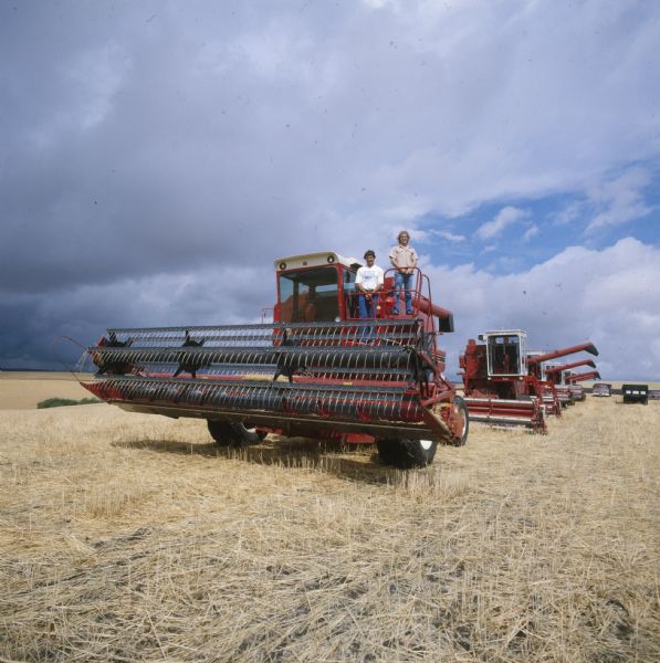 Two men stand posing near the cab of a 1470 Hillside combine, behind them is a row of five combines in a field. There are two trucks and an automobile near the end of the line on the right.