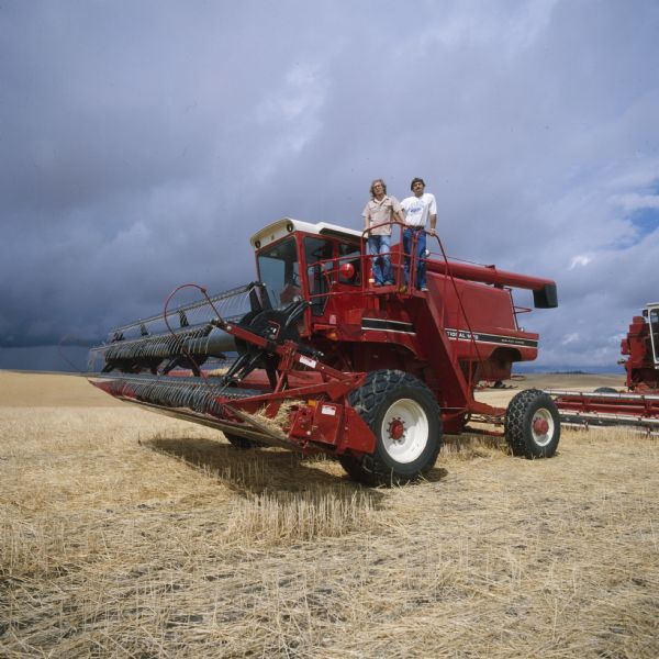 Two men pose on the platform near the cab of a 1470 Hillside combine in a field.