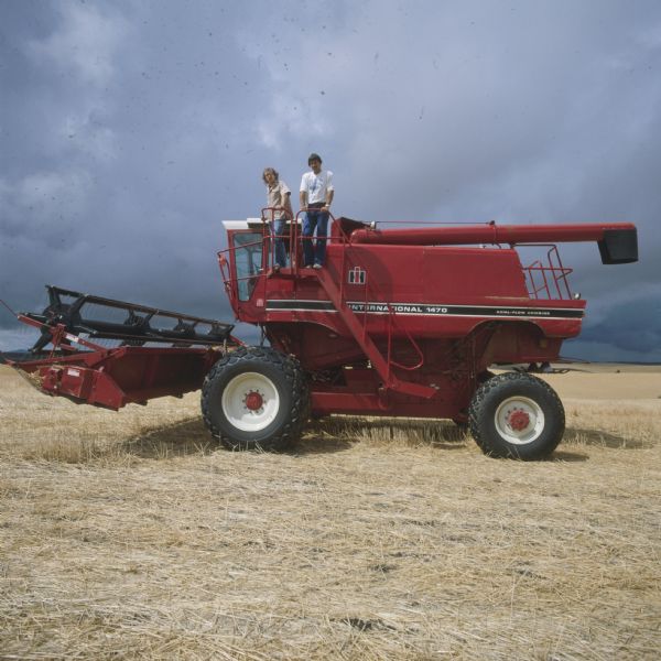 Left side view of two men posing standing on a platform next to the cab of a 1470 Hillside combine in a field.