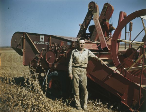 View towards a man wearing tan clothes and a hat standing next to a McCormick combine outdoors in a field.