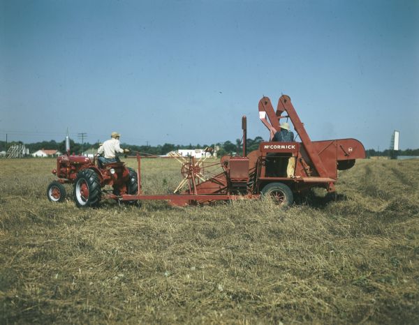 Two men in a field. One is steering a Farmall tractor that is pulling a man on a McCormick combine.