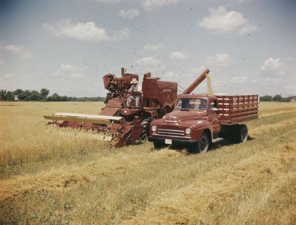 Three-quarter view from left front of a man operating a combine. Next to the combine is a man in a truck. The combine is unloading grain into the bed of the truck.