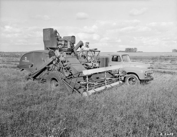 Three-quarter view from right front of a man using a McCormick combine in a field. Parts of the machine have been colorized. There is a man in an International truck parked behind the combine.