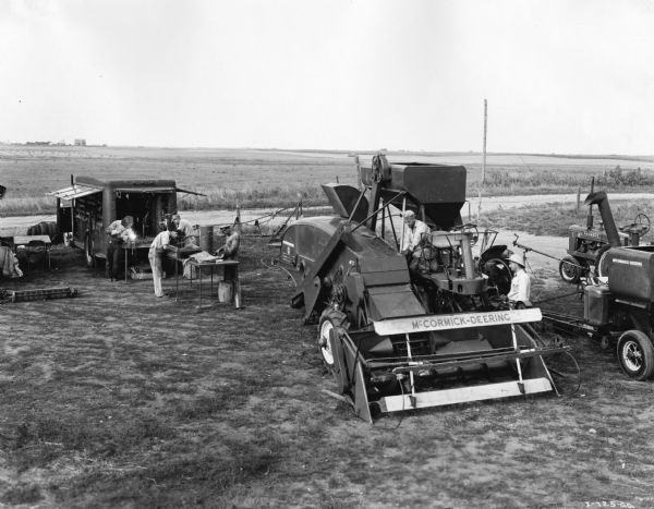Two men near a McCormick-Deering 123 self propelled combine. There are additional men at a table set up near a small trailer. There is a Farmall H is in the background. 