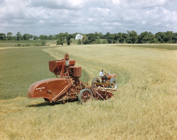 Slightly elevated view from right side of man using a Farmall M tractor to pull a McCormick-Deering 122 combine in a field. Trees and a white frame building are in the far background.
