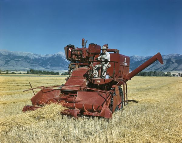 Three-quarter view from front left of a man operating a combine in a field. Mountains are in the background.