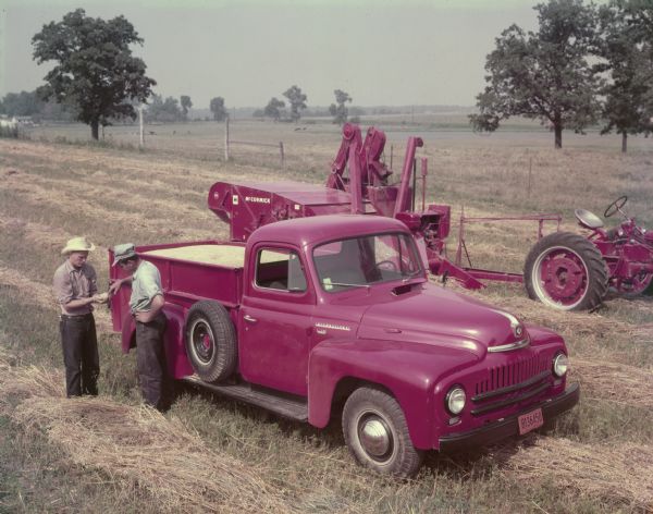 Two men are in a field standing next to an International L0120 truck. Behind the truck is a McCormick 64 combine attached to a tractor.