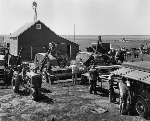 Several men working outside amid agricultural equipment and a trailer. A wood framed building and windmill are in the background. 