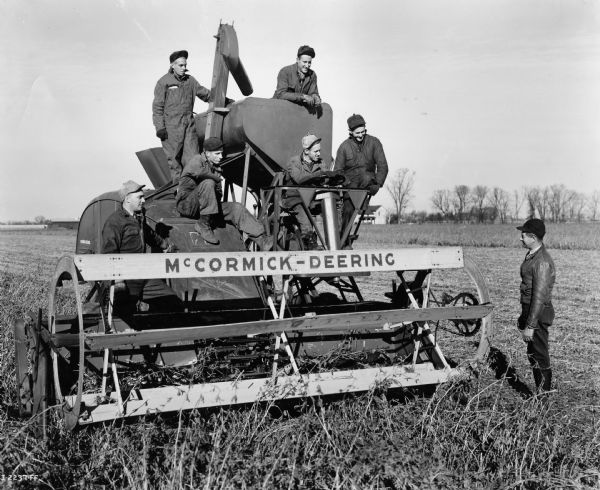 Seven men in overalls and hats are standing on or near an experimental 12 ft self propelled combine in a field.