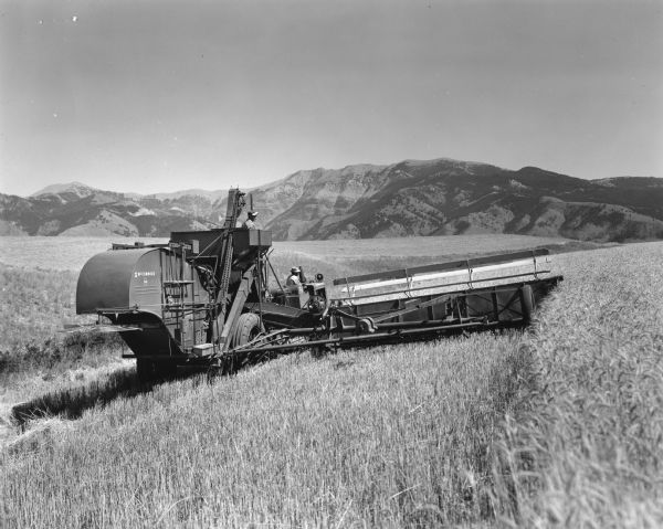 Rear view of a McCormick-Deering No.140 pull type combine being pulled by an International Diesel TD-MA in a field. There are mountains in the distance.