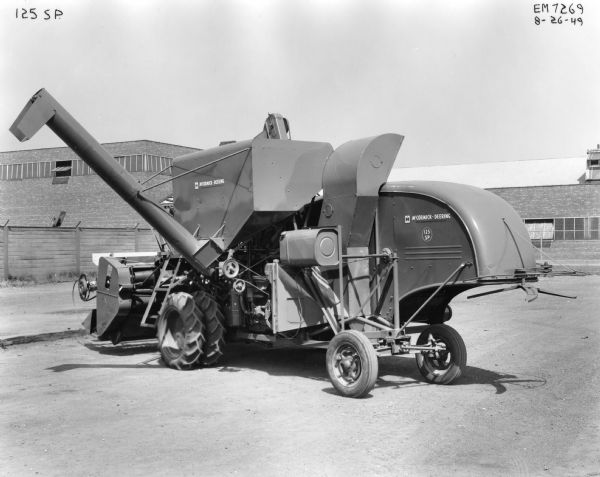 Three-quarter view from behind of a McCormick-Deering no. 125 self propelled combine parked outdoors. There are brick buildings and a wooden fence in the background. 
