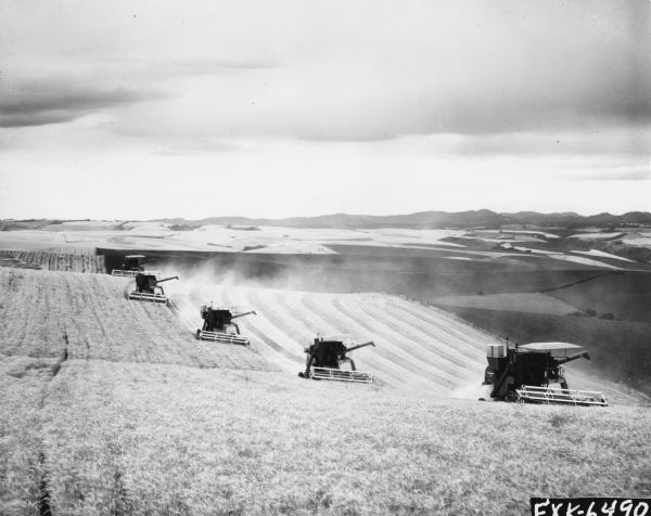 Elevated view of five McCormick-Deering combines harvesting a field with hills. From an advertising series for the McCormick-Deering self-propelled combines models no. 82, 205, 105, 315, 403, and 503.