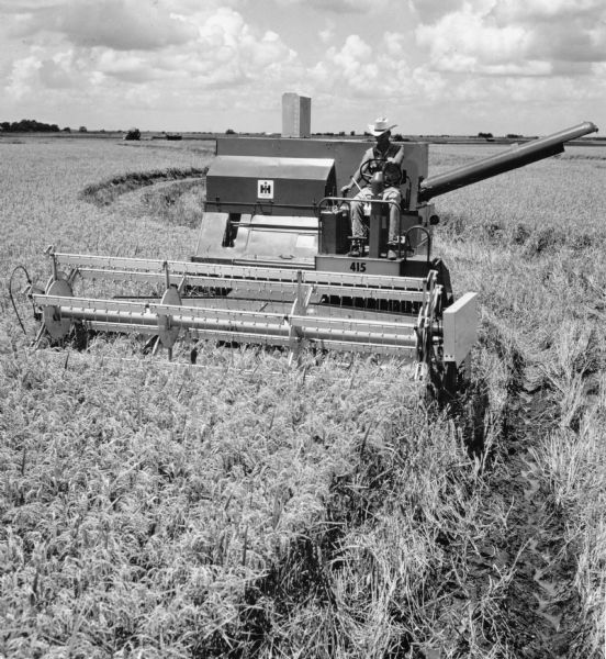 Slightly elevated view from in front of a 415 self propelled combine harvesting rice. The combine is driven by a man in a white cowboy hat, and the IH logo is clearly visible. Photograph selected from an advertising series for the McCormick-Deering self-propelled combines models no.82, 205, 105, 315, 403, and 503.