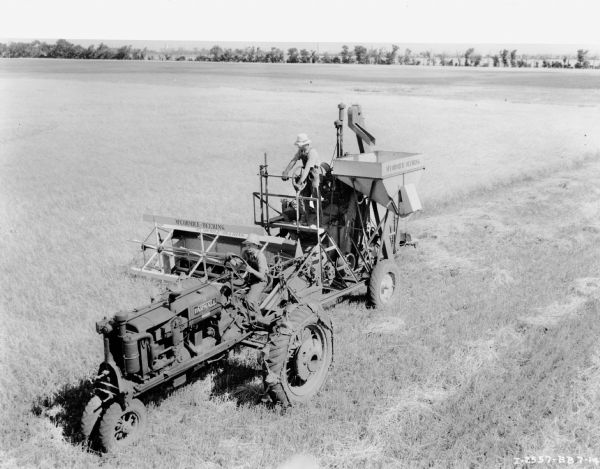 Elevated front left view of a man in a field driving a Farmall F-30 pulling a McCormick-Deering pull type combine. The combine appears to be a No. 80 pull type combine. 
