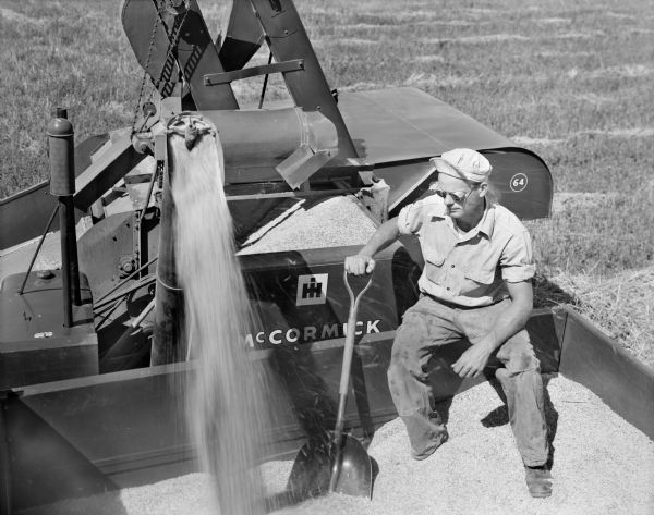 View looking down at a man, wearing safety glasses, sitting on the edge of a wagon holding a shovel. Behind him a McCormick combine 64 is emptying grain into the wagon.