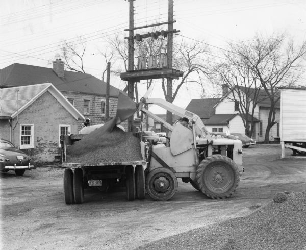 View from the driver's side of a man operating a Hough model HE payloader. He is unloading gravel into a truck. There are buildings and automobiles in the background as well as a large electric transformer.