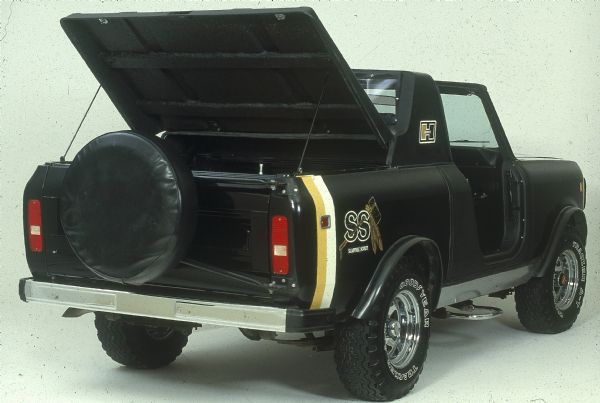 Three-quarter view from rear of passenger side. Passenger side door and cab roof are removed. Spare tire with black cover is attached to rear tailgate. Truck bed cover is in raised position. "Shawnee Scout" decal is at rear of truck bed. "Hurst" decal is behind passenger side door. Goodyear A-T Tracker tires.