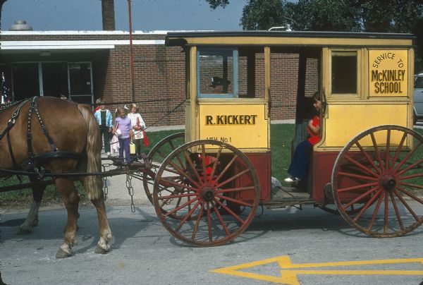 Left side view of Kickert horse-drawn carriage. One child is sitting in the carriage, and behind the carriage a group of children are walking on the sidewalk in front of a school building.